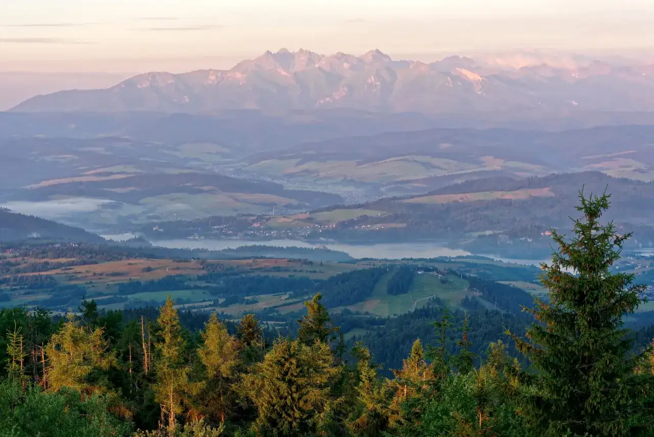 Widok na mgliste doliny i jezioro, a w oddali majestatyczne, oświetlone słońcem gorce szczyty.