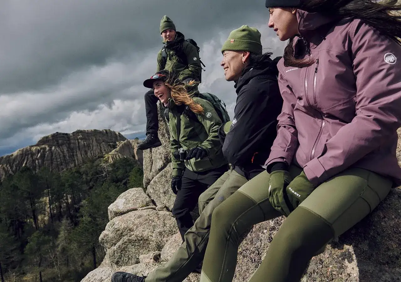 Grupa przyjaci&oacute;ł w kurtkach i spodniach z materiału softshell odpoczywa na skale, podziwiając g&oacute;rski krajobraz.