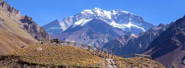 Majestatyczne góry z ośnieżonym szczytem Aconcagua w tle. Ścieżka z drewnianym ogrodzeniem prowadzi przez suchą, trawiastą górę.