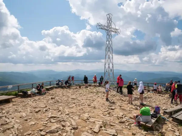 Ludzie odpoczywają na szczycie, podziwiając panoramę Bieszczad. Metalowy krzyż góruje nad kamienistym terenem, świadcząc o popularności tego miejsca na szlaki Bieszczady.