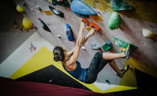Kobieta wspina się po kolorowej ścianie, chwytając się pomarańczowych uchwytów. To właśnie bouldering – sportowa wspinaczka bez lin.