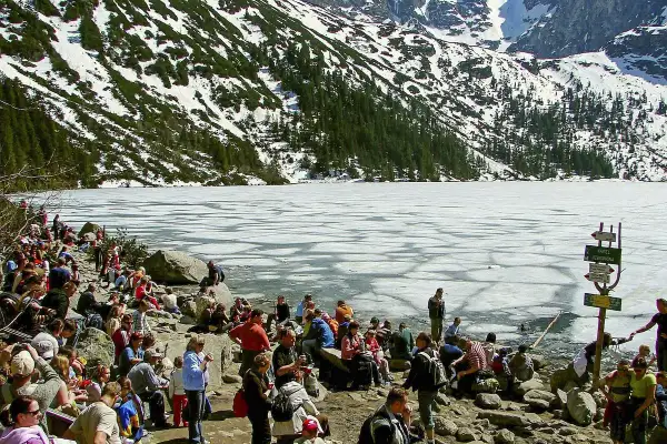 Tłum ludzi odpoczywa nad zamarzniętym jeziorem Morskie Oko, w tle góry pokryte śniegiem i lasem. Palenica Białczańska w oddali.
