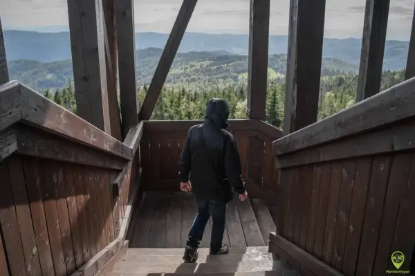 Osoba w czarnej kurtce schodzi po drewnianych schodach wieży widokowej. Przed nią rozpościera się malowniczy widok na górzysty krajobraz, gdzie widać lasy i doliny. To idealne miejsce, by podziwiać piękno przyrody z wysokości, czując chłód górskiego po...
