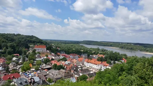 Widok na Kazimierz Dolny z górami, rzeką Wisłą i charakterystyczną zabudową. W oddali widać wzgórza, a na pierwszym planie urokliwe kamienice.