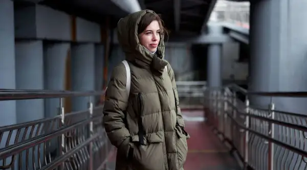 Kobieta w zielonej, pikowanej parkce z kapturem, stojąca na tle miejskiej architektury. Parka co to? To idealna ochrona przed zimnem.