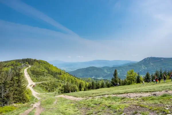 Widok na Beskid Śląski z trasą na Klimczok. W oddali widać szczyt z wieżą. Grupa turystów wędruje po zielonych zboczach.