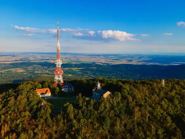 Wieża RTV na górze Ślęża góruje nad lasami i krajobrazem. Obok niej budynki i kościółek.
