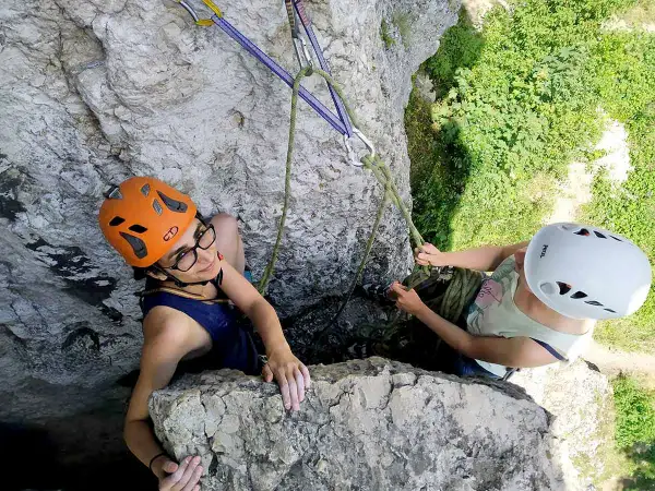 Dwie dziewczyny w kaskach podczas wspinaczki skalnej. Jedna z nich, w pomarańczowym kasku, uśmiecha się do obiektywu, druga, w białym kasku, asekuruje ją.