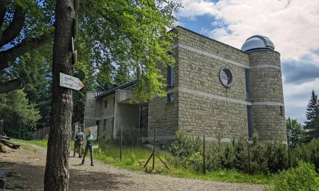 Kamienny budynek obserwatorium astronomicznego na szczycie góry, z kopułą na dachu. Obok tabliczka szlaku turystycznego PTTK, wskazująca drogę do schroniska. W tle las i niebo z chmurami. To miejsce to Lubomir szczyt.